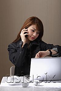 AsiaPix - Young woman sitting at table with lap top, talking on phone, looking at watch