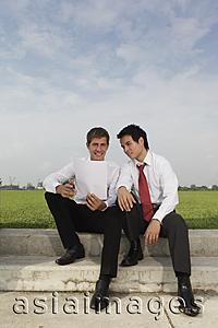 Asia Images Group - Two business men sitting outside looking at piece of paper