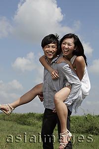 Asia Images Group - young man carrying a woman on his back laughing with blue sky and clouds in background.