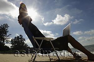 Asia Images Group - Man sitting in beach chair with laptop, sun burst behind his head