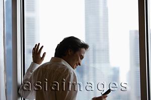 Asia Images Group - Rear view of man standing by window and looking at phone