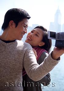 Asia Images Group - Couple looking at each other by waterfront, man holding video camera