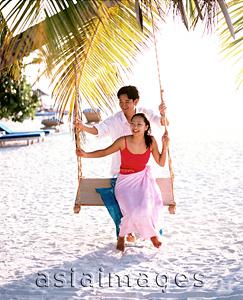 Asia Images Group - Man pushing woman on swing under coconut tree looking off camera, sea in background