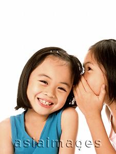 Asia Images Group - Two young girls whispering to each other, laughing, white background.
