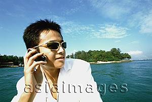 Asia Images Group - Young man with sunglasses using cellular phone, blue sky and water in background