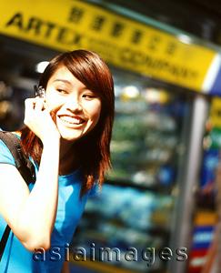 Asia Images Group - Young woman using cellular phone, shops in background.
