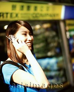 Asia Images Group - Young woman using cellular phone, shops in background.