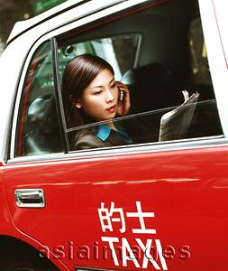 Asia Images Group - Executive woman using cellular phone and reading newspaper in taxi.