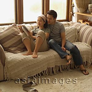 Asia Images Group - Couple sitting on sofa, looking at magazine
