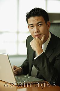 Asia Images Group - Young man with hand on chin, looking at camera, laptop open in front of him