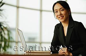 Asia Images Group - Young woman sitting at desk, looking at camera, laptop open next to her
