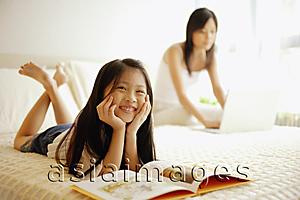 Asia Images Group - Mother using laptop, daughter lying down with book open in front of her