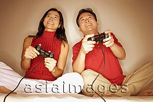 Asia Images Group - Man and woman sitting side by side, legs crossed, playing with handheld video game