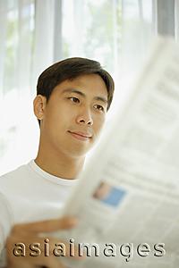 Asia Images Group - Young man reading newspaper, focus on the background