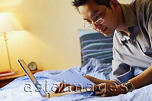 Asia Images Group - Young man on bed using laptop and reading document
