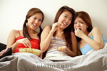 Asia Images Group -  Young women sitting side by side, eating popcorn and watching TV