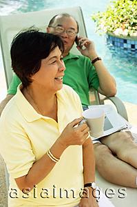 Asia Images Group - Woman holding coffee mug, man on lounge chair, using mobile phone