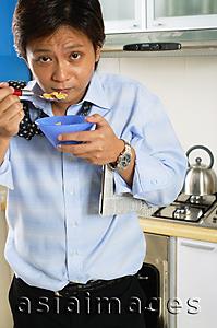 Asia Images Group - Man in kitchen, eating bowl of cereal, looking at camera