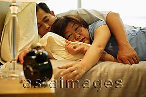 Asia Images Group - Mature couple sleeping in bedroom, woman reaching for alarm clock