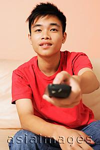 Asia Images Group - Teenage boy sitting on sofa, pointing remote control towards camera