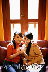 Asia Images Group - Young women sitting in living room, one holding cup, looking at camera