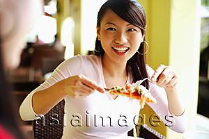 Asia Images Group - Young woman lifting slice of pizza with cutlery, smiling at another person in front of her