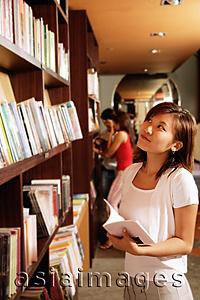Asia Images Group - Young woman at bookstore, holding book, looking up