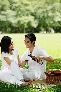 Asia Images Group - Couple sitting with picnic basket, smiling at each other, man pouring wine from bottle