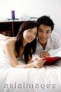 Asia Images Group - Couple lying on bed, holding book, looking at camera