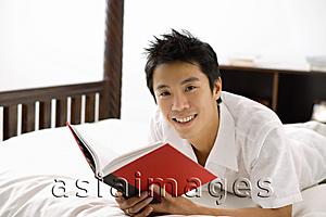 Asia Images Group - Man lying on bed, reading book, smiling at camera