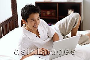 Asia Images Group - Man lying on bed, using laptop, smiling