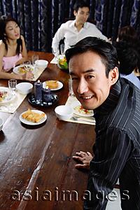 Asia Images Group - Man leaning on table, smiling at camera, people in the background