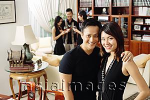 Asia Images Group - Couple in living room, smiling at camera, people in the background