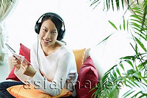 Asia Images Group - Woman sitting on sofa, wearing headphones, holding CD case, smiling