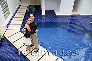 Asia Images Group - Couple standing by swimming pool, embracing, looking up at camera