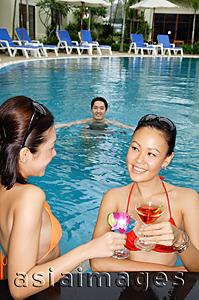 Asia Images Group - Young women talking in swimming pool, holding cocktails, man swimming behind them
