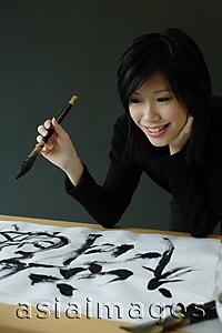 Asia Images Group - Woman holding paintbrush, looking at Chinese script on paper