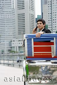 Asia Images Group - Businessman sitting in trishaw, using mobile phone, buildings in the background