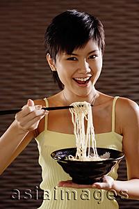 Asia Images Group - Young woman lifting noodles from bowl with chopsticks, smiling at camera