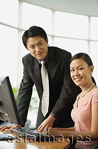 Asia Images Group - Male executive and female executive by desk in office, smiling at camera