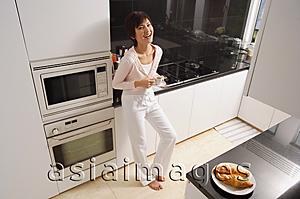 Asia Images Group - Young woman standing in kitchen, holding cup and saucer, smiling at camera