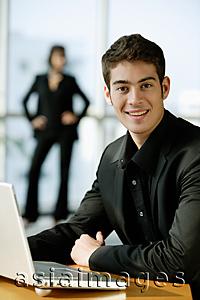 Asia Images Group - Male executive dressed in black, sitting next to laptop, looking at camera, smiling