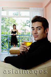 Asia Images Group - Man sitting in living room, holding a glass of wine, woman in the background
