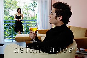 Asia Images Group - Man sitting in living room with a glass of wine, woman standing in the balcony