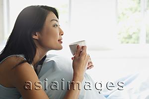 Asia Images Group - Woman sitting on bed, holding cup, looking away