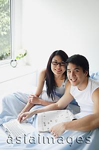 Asia Images Group - Couple sitting side by side in bedroom with laptop, smiling at camera