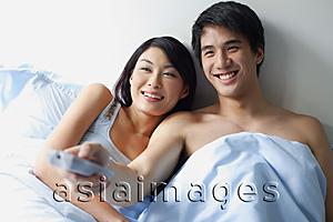 Asia Images Group - Couple sitting up in bed, watching TV, man holding TV remote control