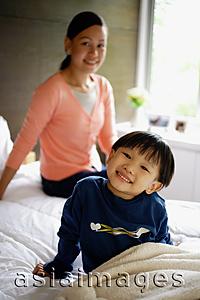 Asia Images Group - Young boy sitting on bed, smiling at camera, mother in the background
