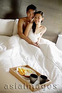 Asia Images Group - Couple sitting up in bed, looking at camera, breakfast tray at foot of bed