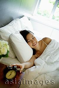 Asia Images Group - Woman reaching for alarm clock on bedside table, smiling